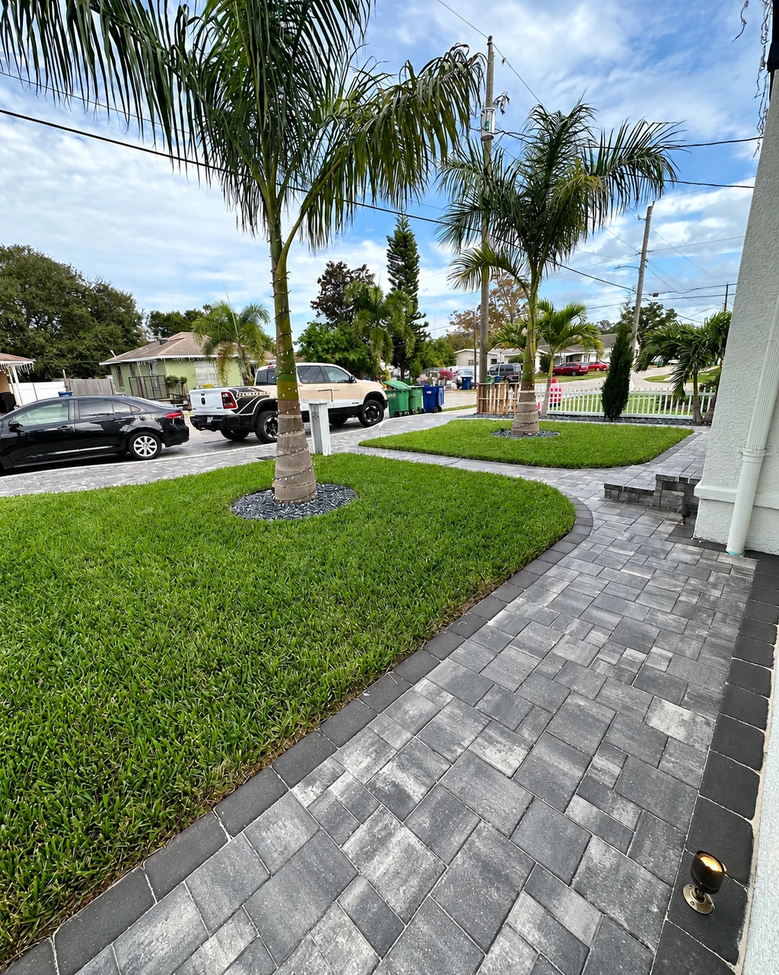 Gray paver garden path follows a curved lawn edge beside palms, landscape lighting and the white house.